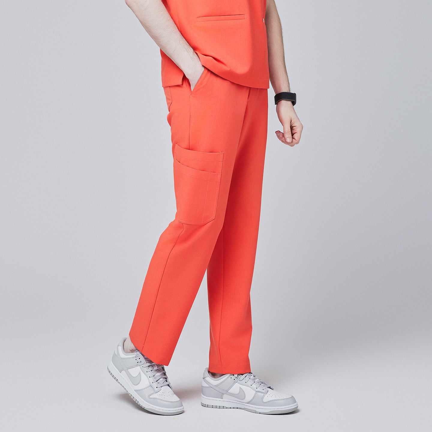 Image of Angled side view of tiger orange scrub pants with spacious side pocket detailing and a tapered fit, paired with light gray sneakers, modeled against a neutral gray background,Tiger Orange